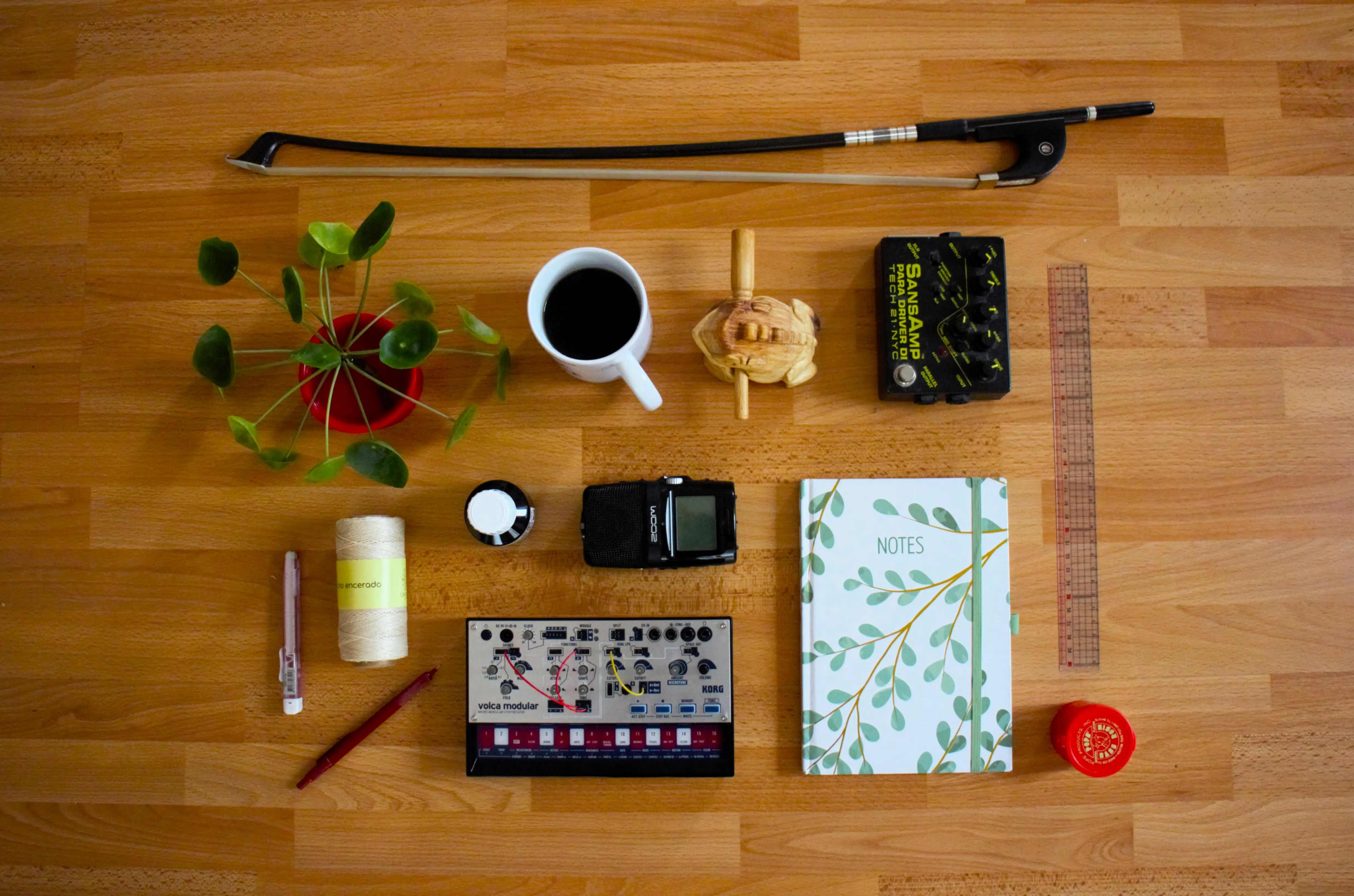 An image of several objects aligned in an organised way. You can see a double bass bow, a plant sprout, a coffee cup, a percussion frog, an electric bass effect, a pencil, string, ink, a recording device, a synthesizer, a notebook, a ruler and rosin.